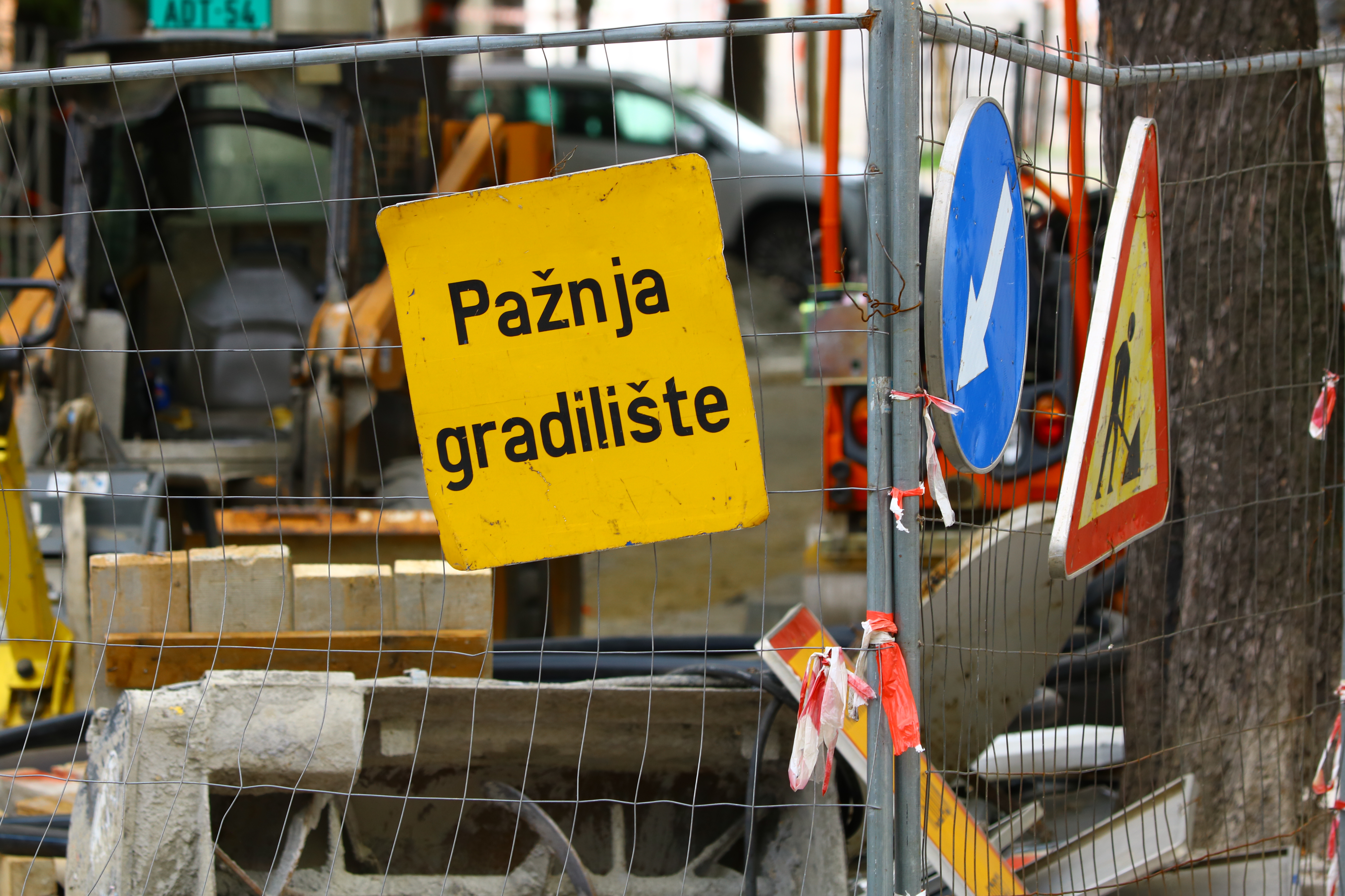 Žuti znak „Pažnja gradilište" na zaštitnoj ogradi gradilišta, pored saobraćajnih znakova i građevinske mehanizacije