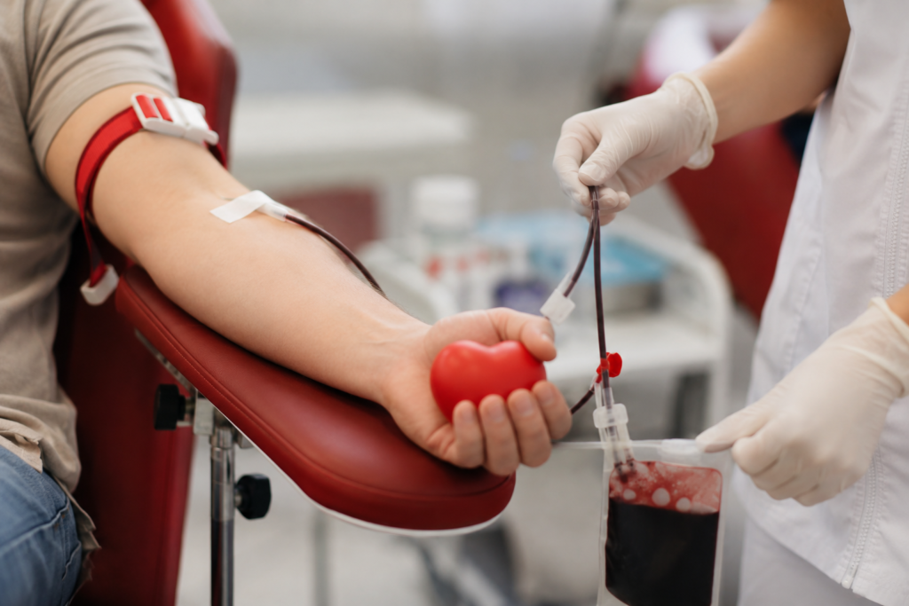 Osoba daje krv u transfuzijskoj stolici, u ruci drži crveno srce, medicinska sestra priključuje kesu za krv