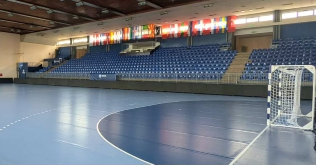 Prazan rukometni teren u sportskoj hali sa plavim tribinama i zastavama evropskih zemalja iznad njih