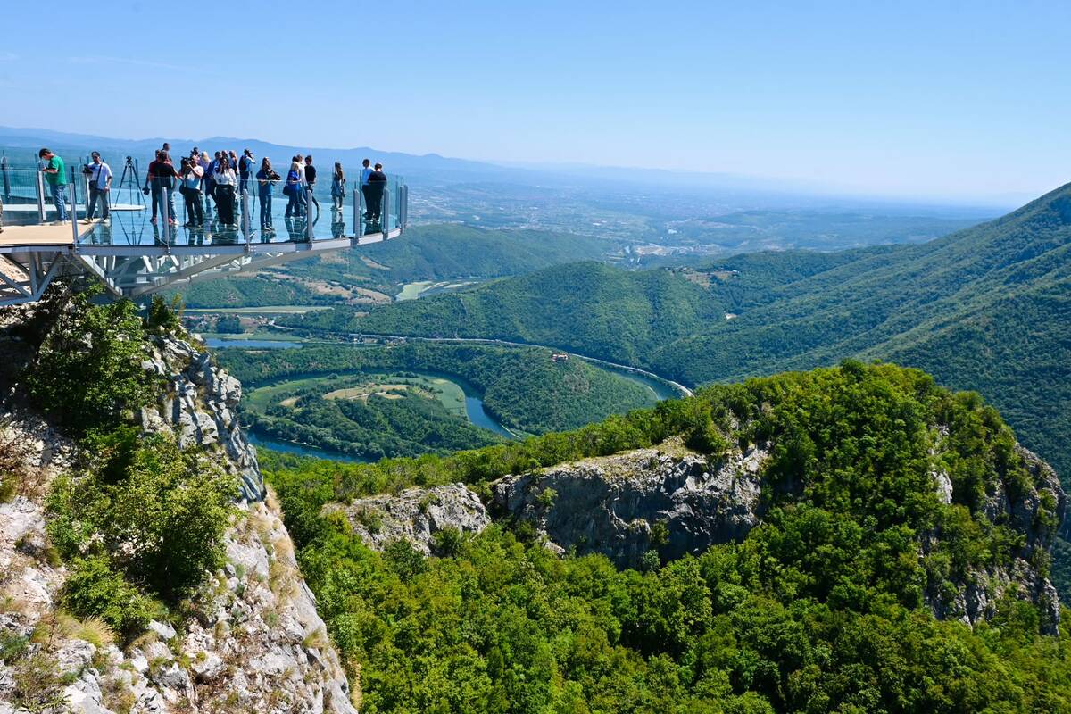 Grupa posetilaca na staklenoj platformi vidikovca iznad zelene klisure, u dubini meandar reke i planinski masiv