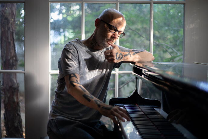 Moby sedi za klavirom pored velikog prozora, naslonjen na instrument, sa naočarima i tetovažama na rukama