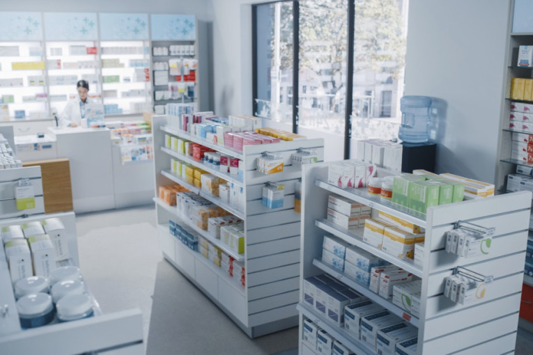 Unutrašnjost apoteke sa policama punim kutija lekova i farmaceutkinjom za pultom