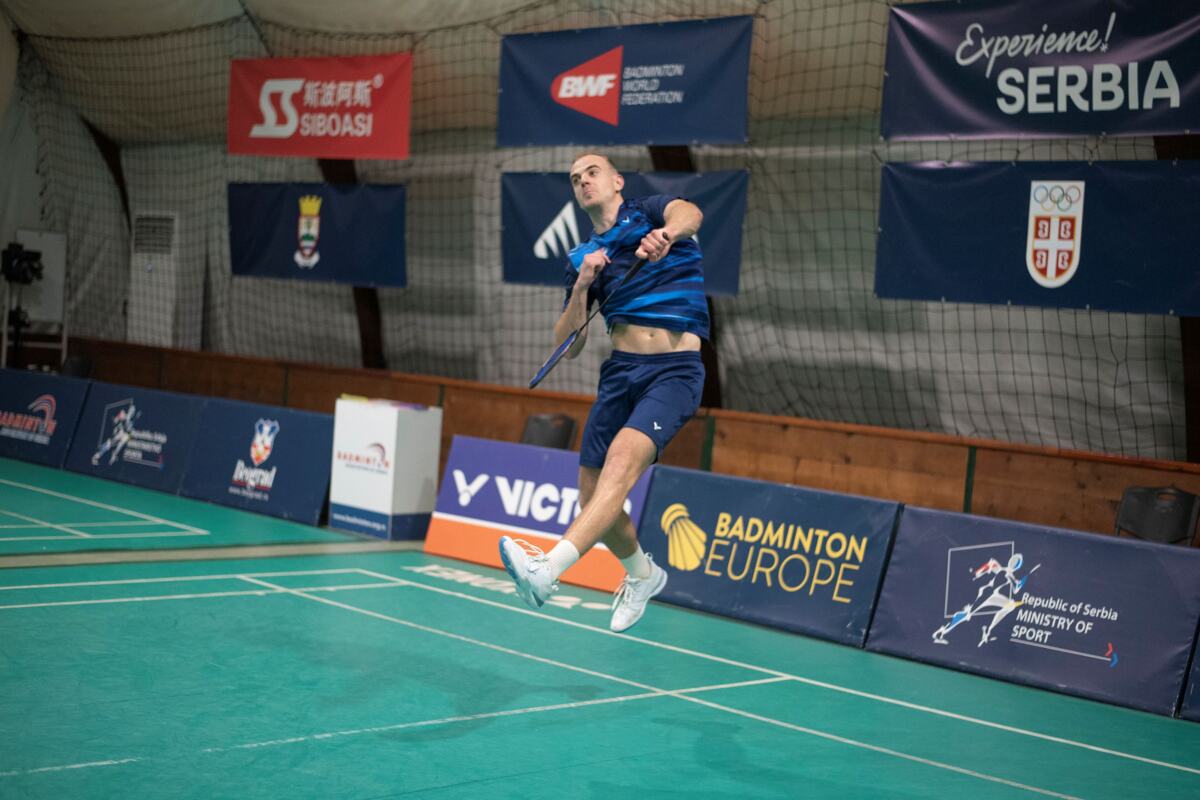 Badmintonista u plavom dresu u skoku izvodi smeč na terenu sa banerima Badminton Europe i Badminton World Federation