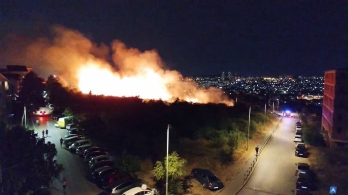 Noćni snimak velikog požara na zelenoj površini pored stambenih zgrada, sa vatrogasnim i hitnim vozilima na prilazu