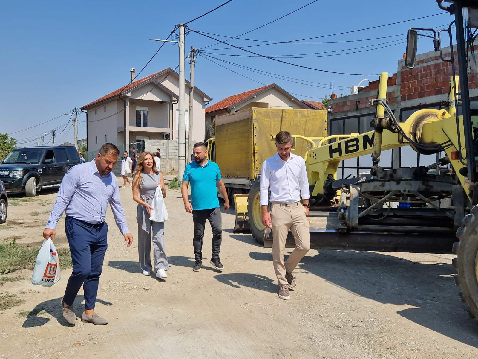 Opštinski funkcioneri i meštani hodaju neasfaltiranom ulicom pored žute građevinske mašine i kamiona u Jajincima