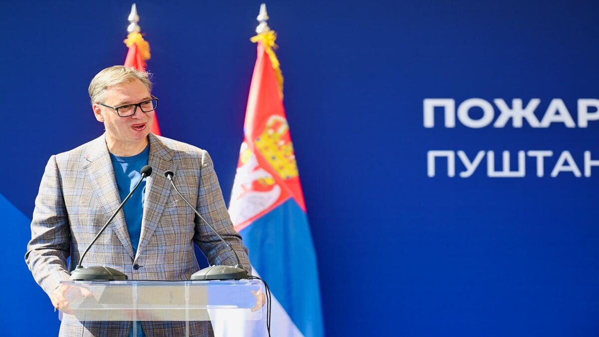 Aleksandar Vučić govori za providnim pultom ispred plave pozadine, iza njega dve zastave Srbije