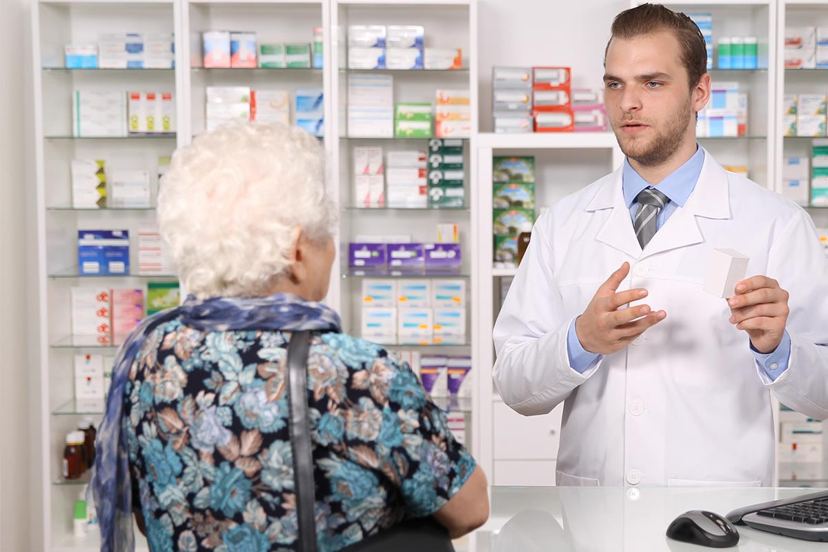 Farmaceut u beloj kecelji predaje kutiju leka starijoj ženi za apotekarskim pultom