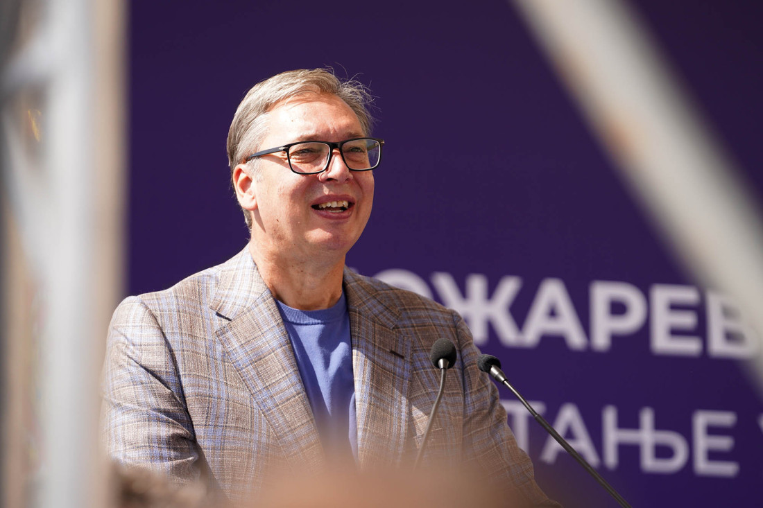 Aleksandar Vučić se osmehuje dok govori na mikrofon ispred ljubičaste pozadine