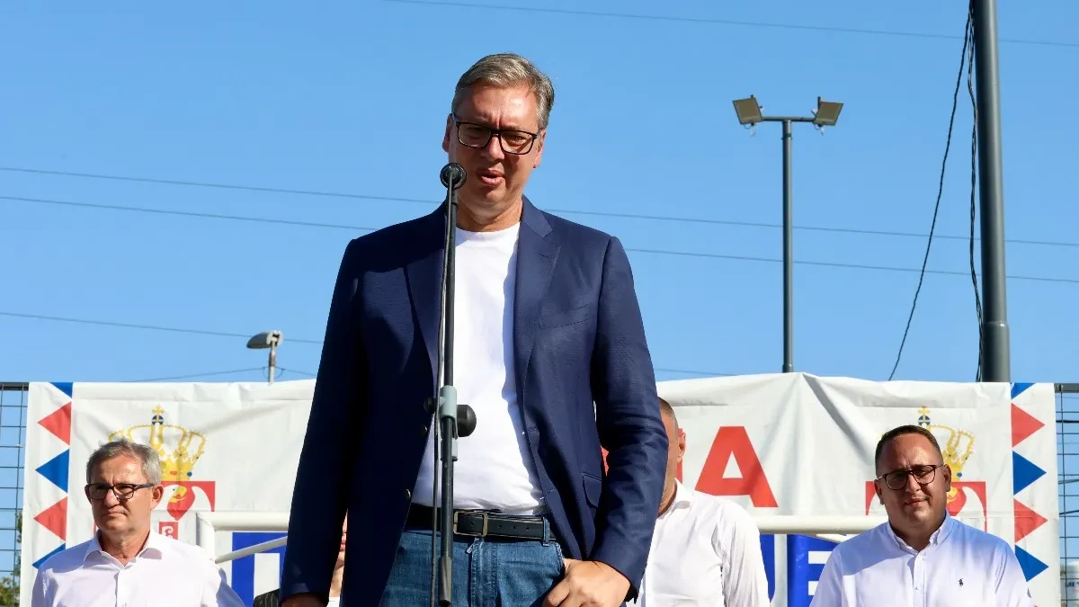 Aleksandar Vučić govori za mikrofonom na otvorenom, iza njega saradnici i bela zastava