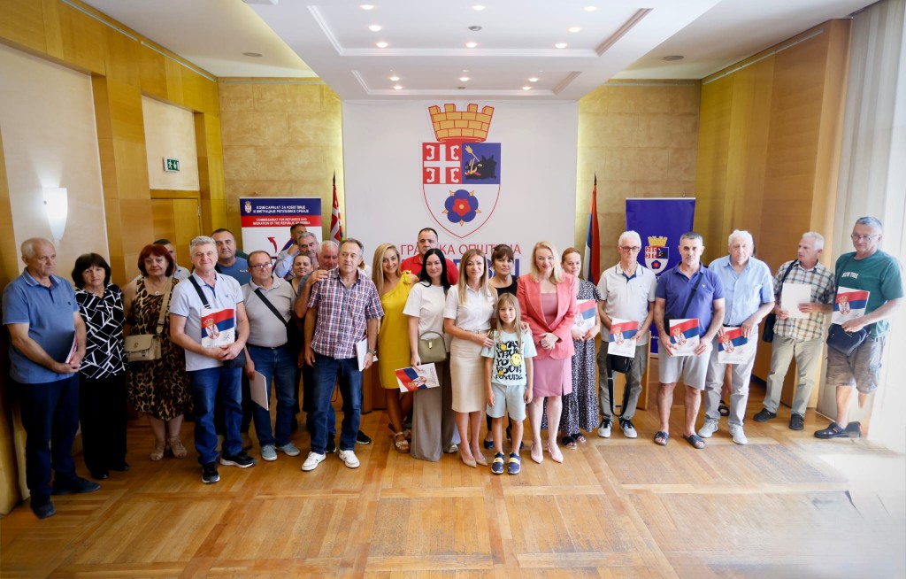 Grupna fotografija predstavnika opštine Voždovac i porodica izbeglica i raseljenih lica u sali opštine, sa grbom Beograda u pozadini