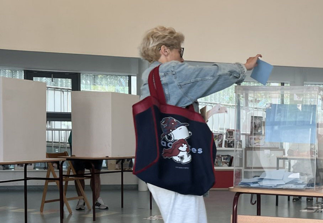 Biračica ubacuje listić u providnu glasačku kutiju, u pozadini kartonski paravani