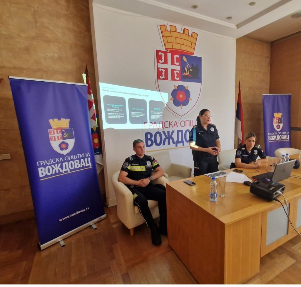 Policijski službenici drže predavanje u svečanoj sali opštine Voždovac, iza njih projekcija prezentacije i baneri Gradske opštine Voždovac