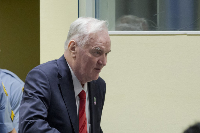 Ratko Mladić u tamnom odelu sa crvenom kravatom u sudnici, pored njega pripadnik obezbeđenja
