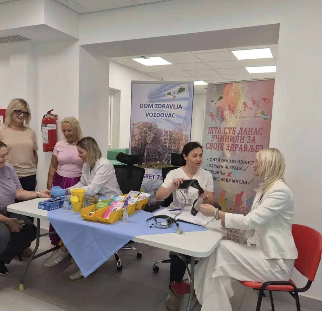 Zdravstveni radnici mere krvni pritisak i pregledaju građane za stolom u zdravstvenoj stanici, iza njih baner Doma zdravlja Voždovac