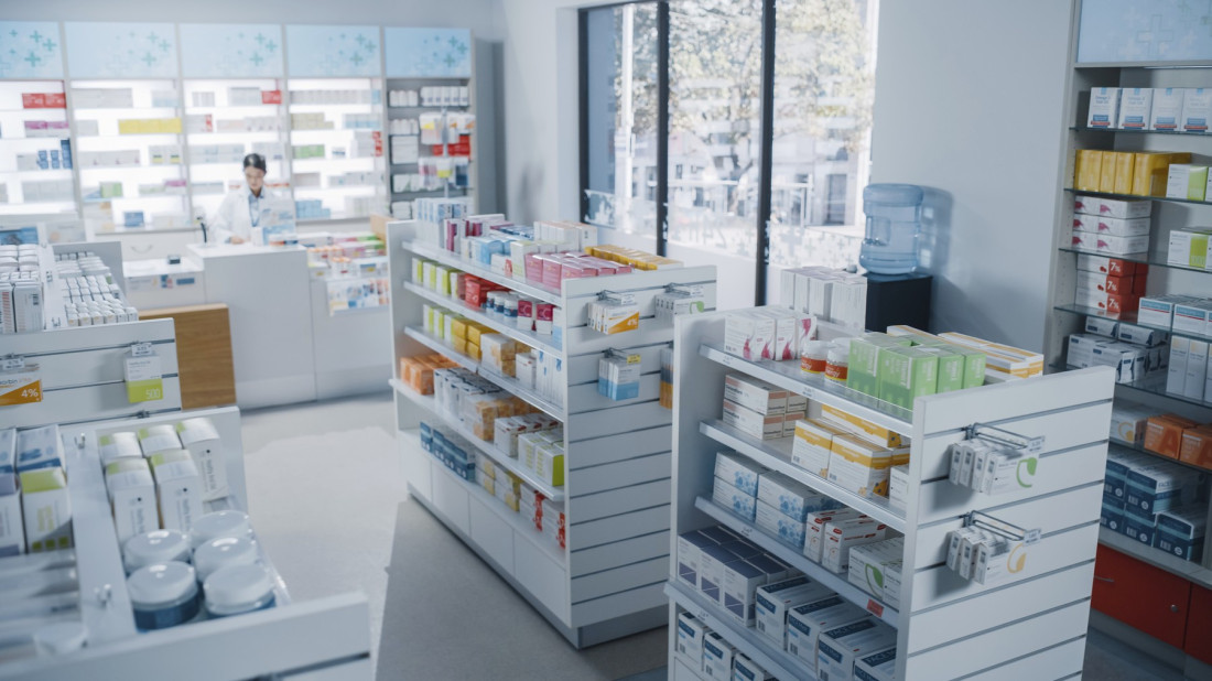 Unutrašnjost apoteke sa policama punim kutija lekova i farmaceutkinjom za pultom u pozadini
