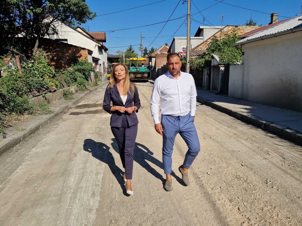 Predsednica opštine i saradnik hodaju sredinom raskopane ulice, u pozadini valjak za asfaltiranje
