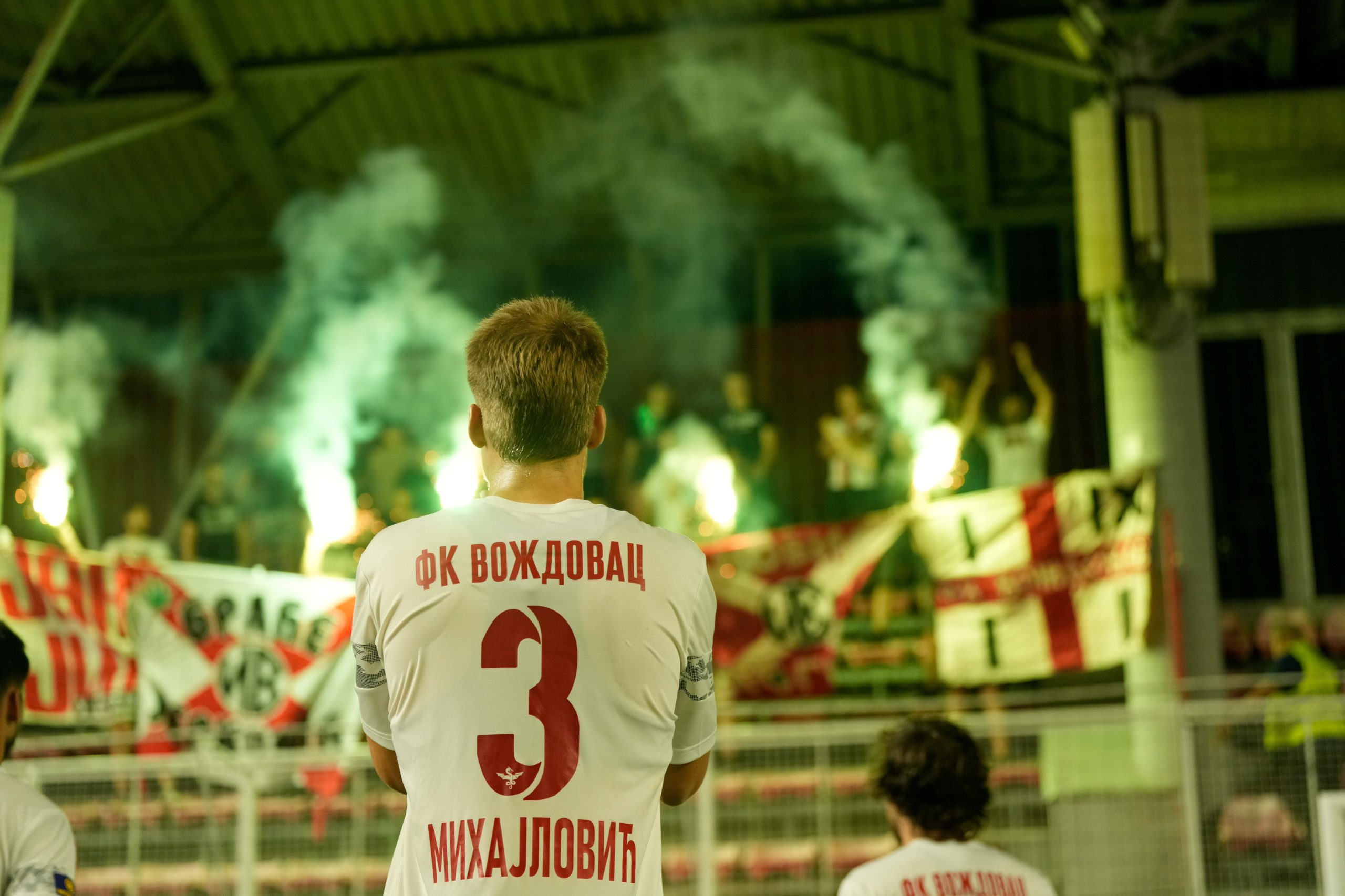 Fudbaler Voždovca sa brojem 3 na dresu gleda ka navijačima koji pale zelene baklje iza ograde tribine