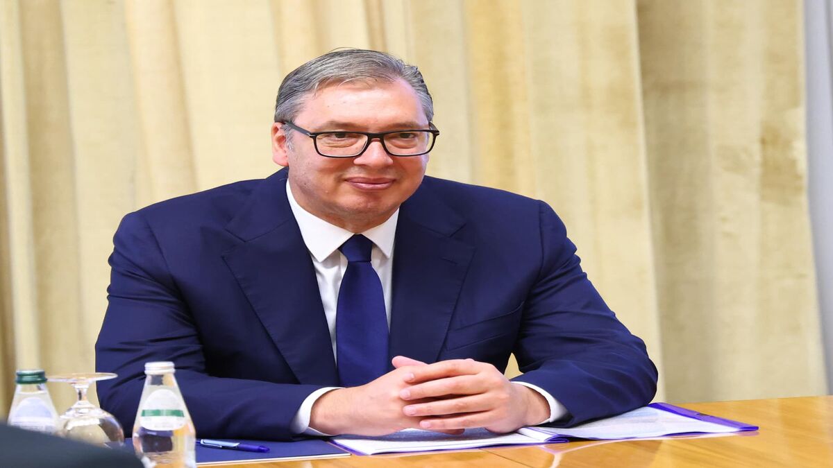 Aleksandar Vučić za stolom u odelu, sklopljenih ruku, ispred zavese