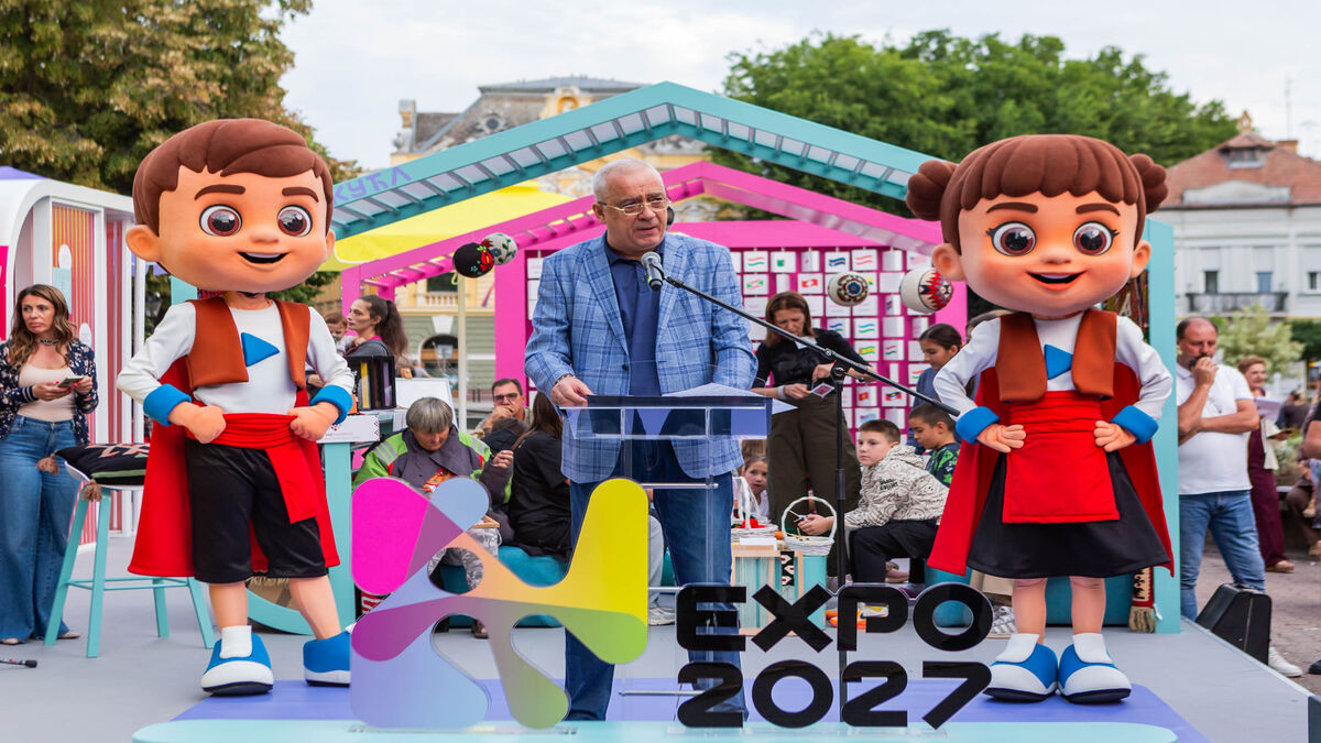 Govornik za pultom na bini izmedju dve maskote EXPO 2027, ispred publike na otvorenom