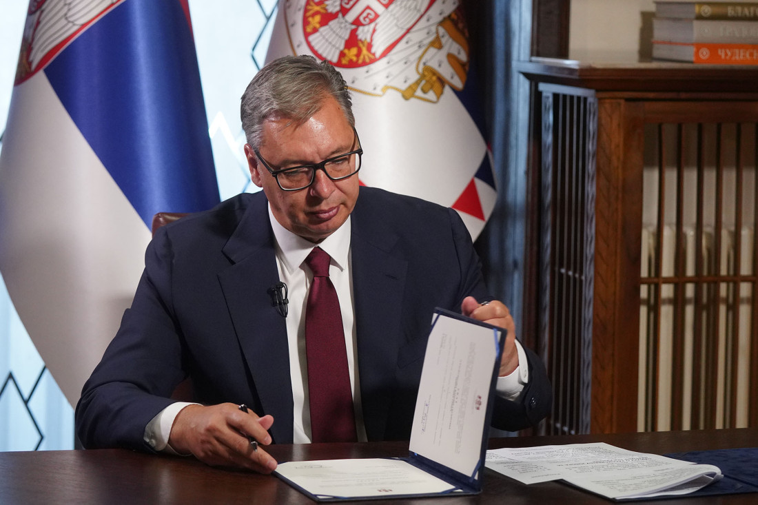 Aleksandar Vučić za stolom potpisuje dokument, iza njega zastave Srbije
