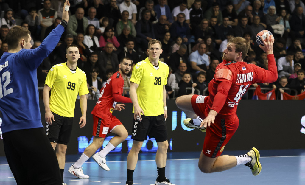 Rukometaš Srbije u crvenom dresu u skoku šutira na gol, ispred njega igrači u žutim dresovima i golman u plavom
