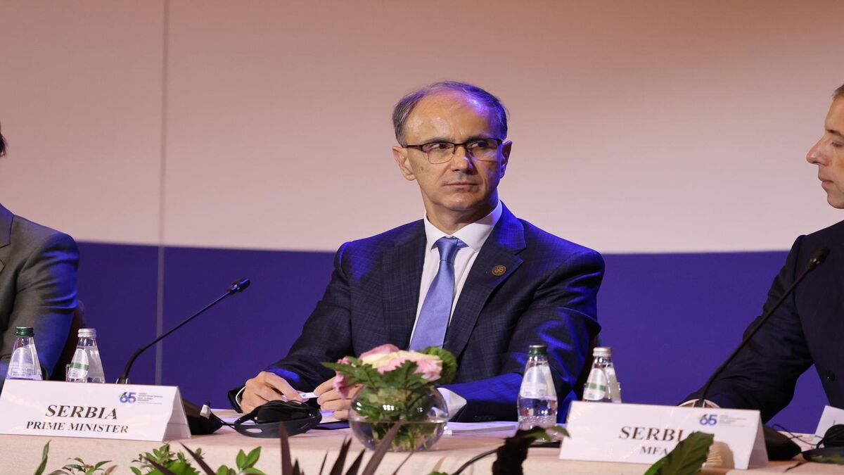 Đuro Macut sedi za stolom na skupu, ispred njega tablica sa natpisom Serbia Prime Minister