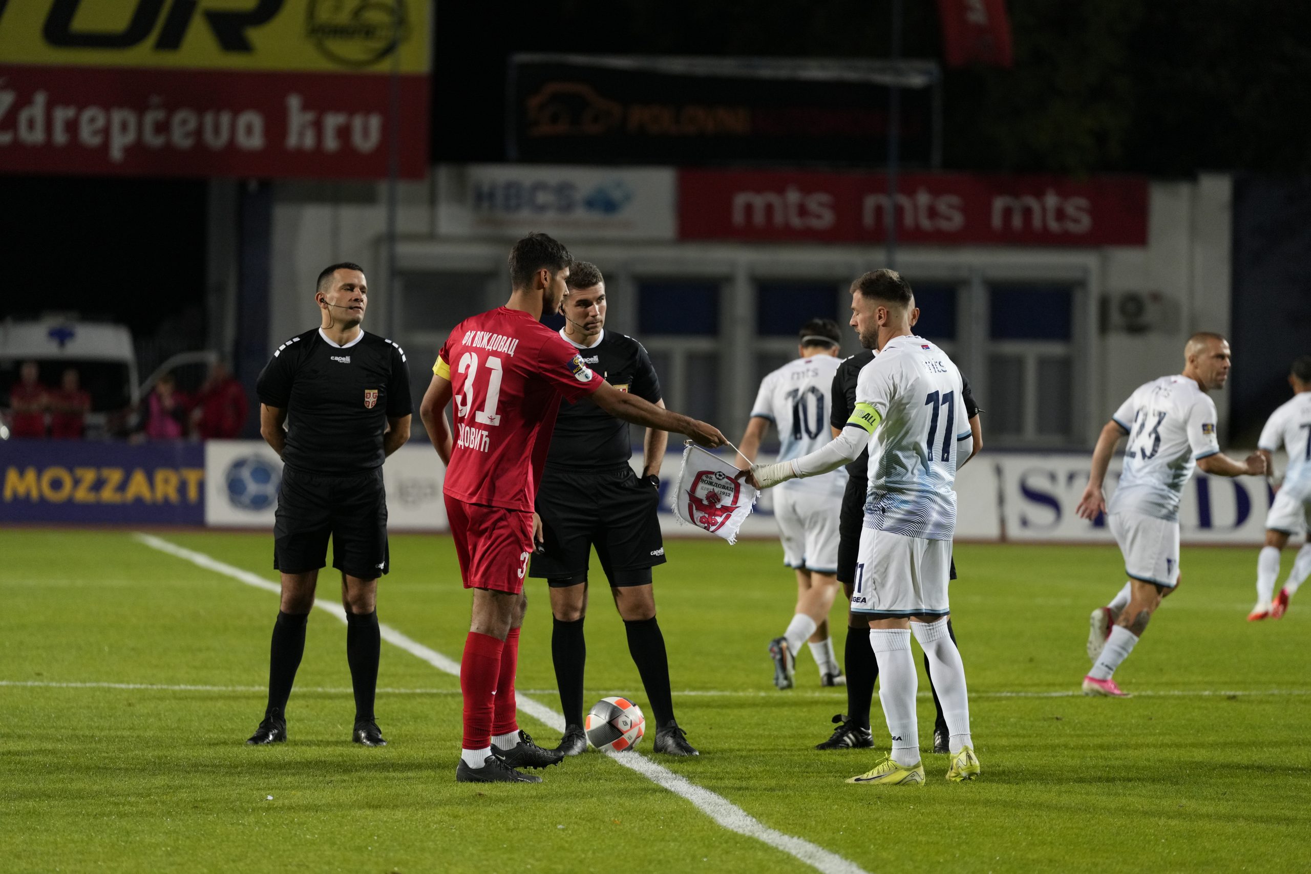 Kapiten FK Voždovac u crvenom dresu i kapiten protivnika u belom razmenjuju zastavice pred početak utakmice, pored sudija