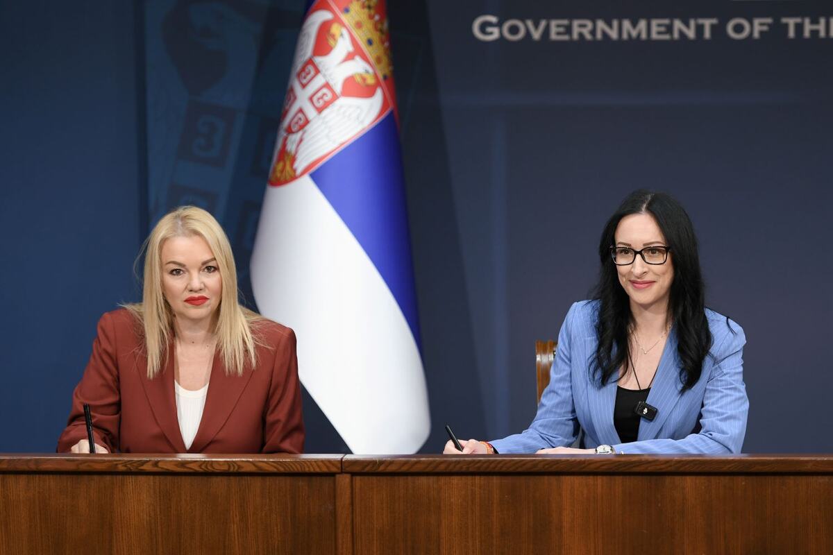 Jelena Žarić Kovačević i Nataša Stanisavljević sede za stolom i potpisuju dokument ispred zastave Srbije