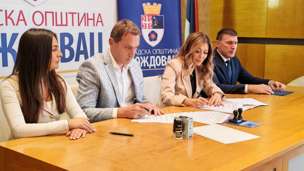 Potpisivanje ugovora za stolom ispred panoa Gradske opštine Voždovac, četvoro učesnika potpisuje dokumente