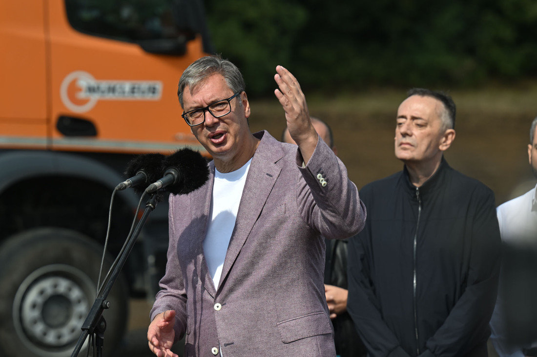 Aleksandar Vučić govori za mikrofonima na otvorenom, iza njega narandžasti kamion