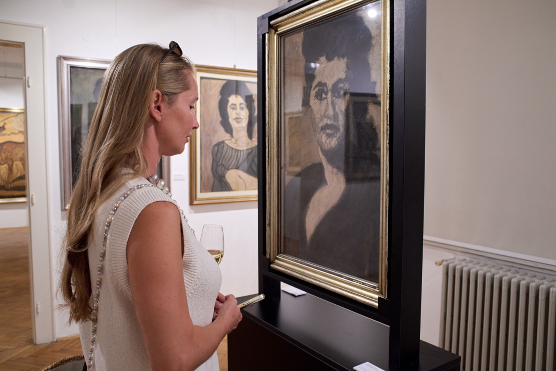 Posetiteljka razgleda uramljeni portret žene na postolju, u pozadini slike na zidu galerije