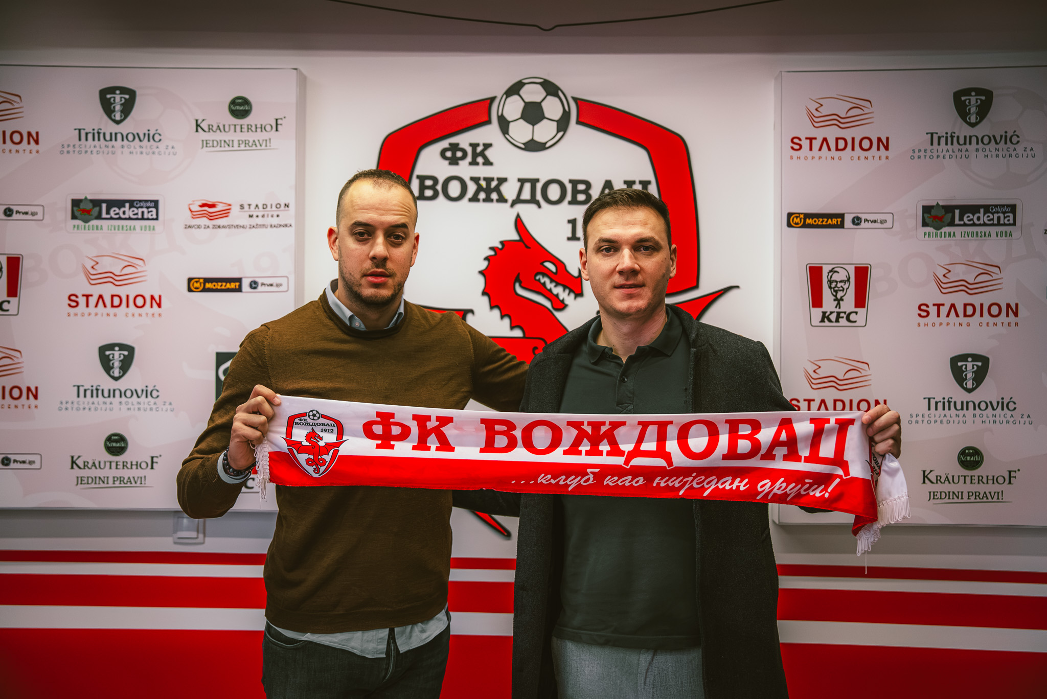 Dvojica muškaraca drže navijački šal FK Voždovac ispred zida sa grbom kluba
