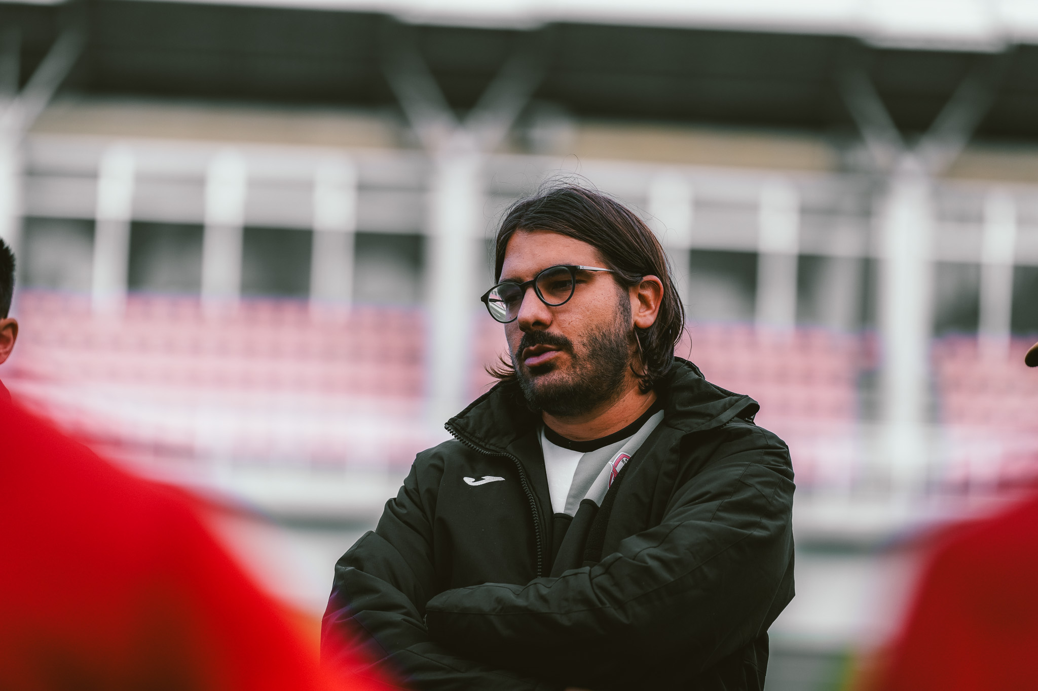 Trener u crnoj jakni sa naočarima na terenu, u pozadini tribine stadiona