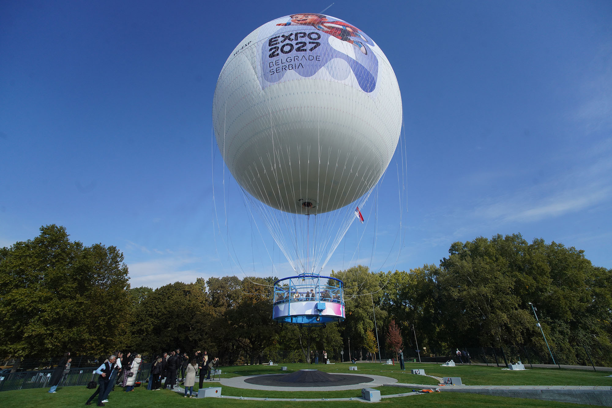 Beli panoramski balon sa natpisom Expo 2027 Belgrade Serbia lebdi iznad travnjaka na Adi Ciganliji, ispod se okupljaju posetioci