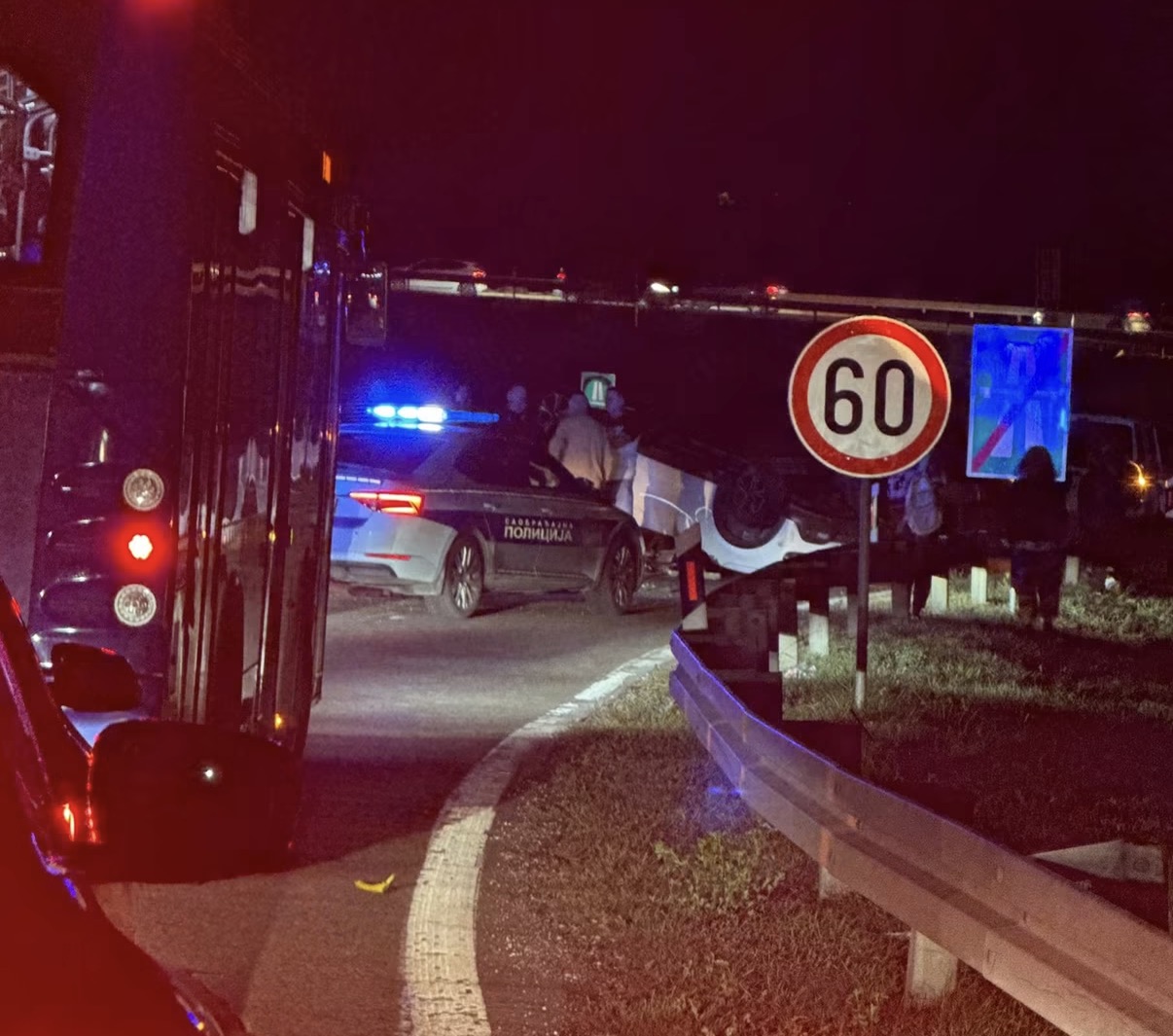 Prevrnut beli automobil pored puta noću, uz vozilo saobraćajne policije i znak ograničenja brzine 60